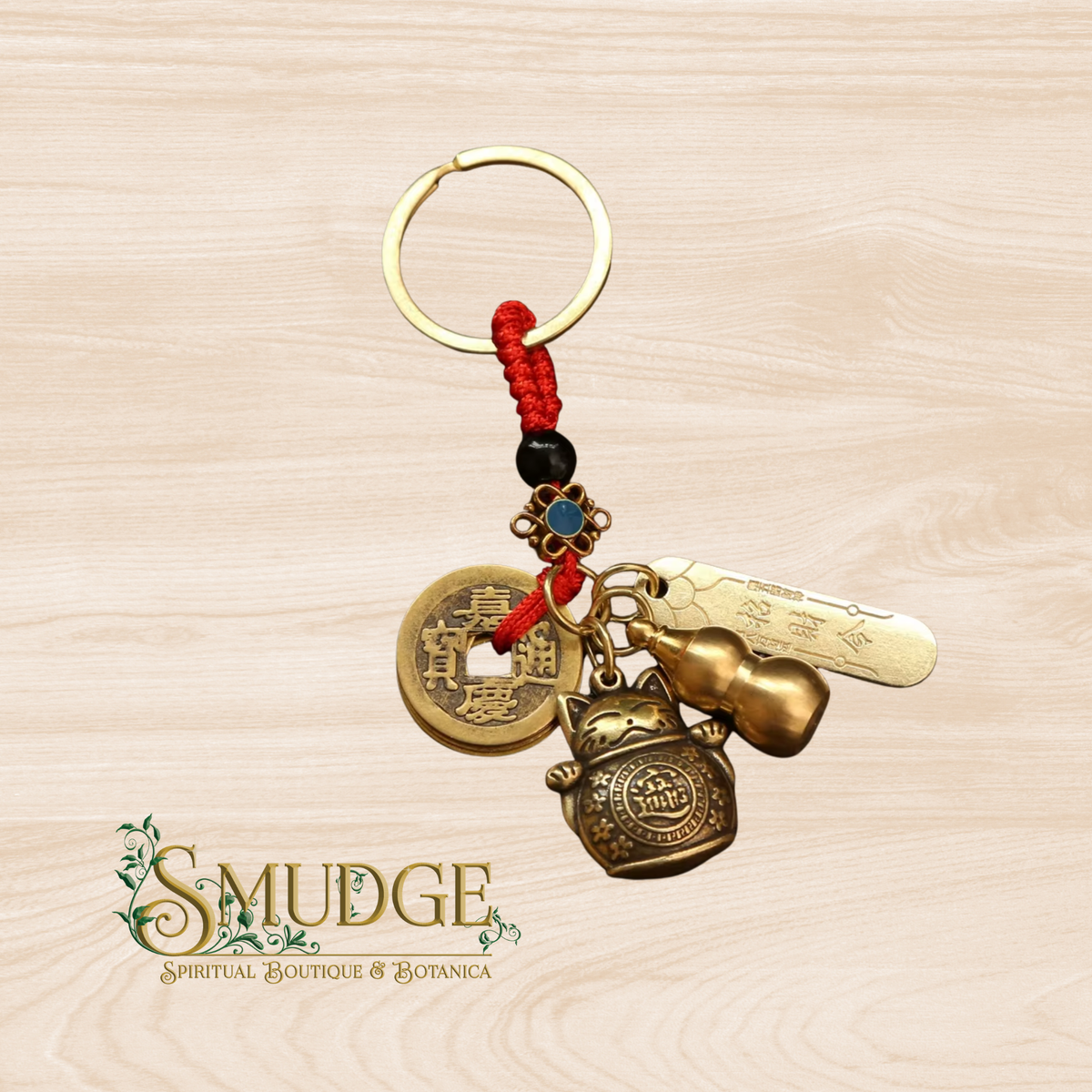 A brass keychain featuring a red braided cord, a gold split ring, a Chinese coin charm, a lucky cat charm, a gourd charm, and a engraved brass tag, displayed on a light wood background with the 'SMUDGE' brand logo in the lower-left corner.