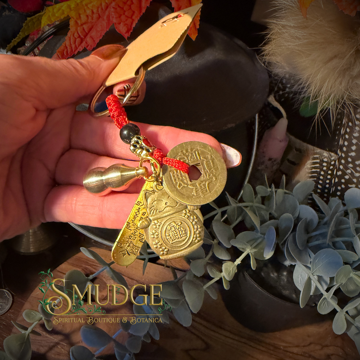 A hand holds a brass keychain with a red cord, featuring a Chinese-style coin, a small bell, and a wooden tag. The background contains decorative plants and a dark container, with a 'Smudge Spiritual Boutique & Botanica' watermark at the bottom.