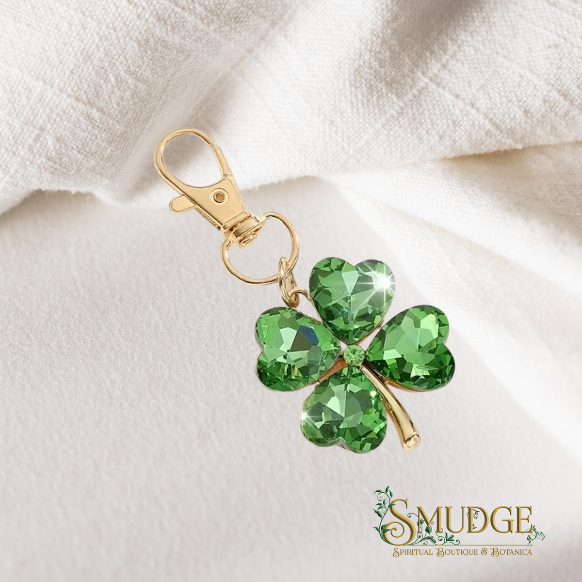 A gold-tone keychain or bag charm shaped like a four-leaf clover with faceted green crystal stones, resting on a textured white fabric background. The brand name 'SMUDGE Spiritual Goods & Botany' is visible in the bottom right corner.