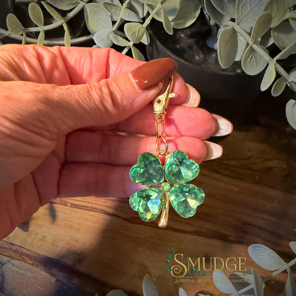 A hand holds a gold-tone St. Patrick's Day charm shaped like a four-leaf clover made of faceted green gemstones, with a lobster clasp at the top.