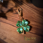 A green faceted crystal four-leaf clover charm with a gold-tone lobster clasp, resting on a dark wooden surface with a 'SMUDGE' brand logo in the bottom right corner.
