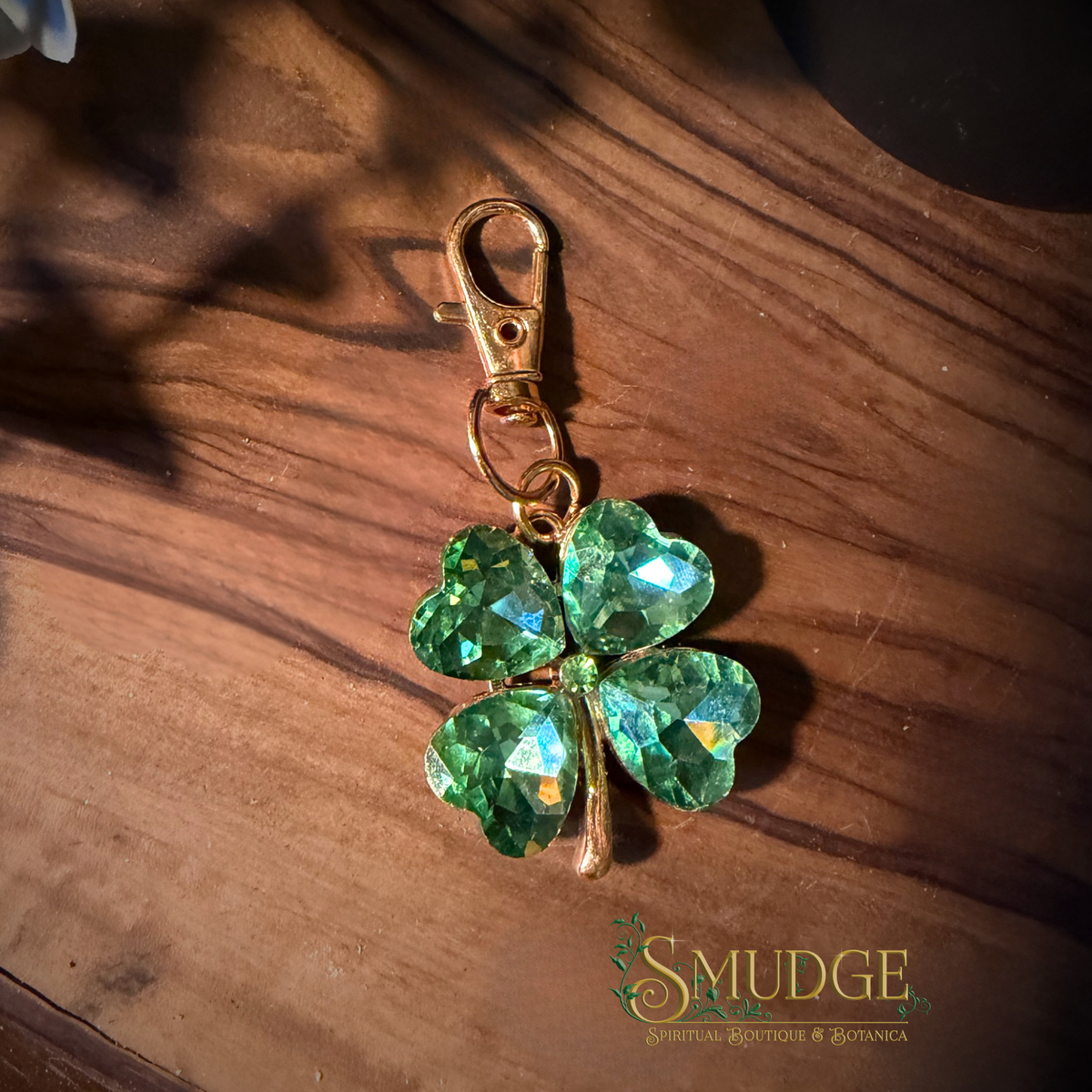 A green faceted crystal four-leaf clover charm with a gold-tone lobster clasp, resting on a dark wooden surface with a 'SMUDGE' brand logo in the bottom right corner.