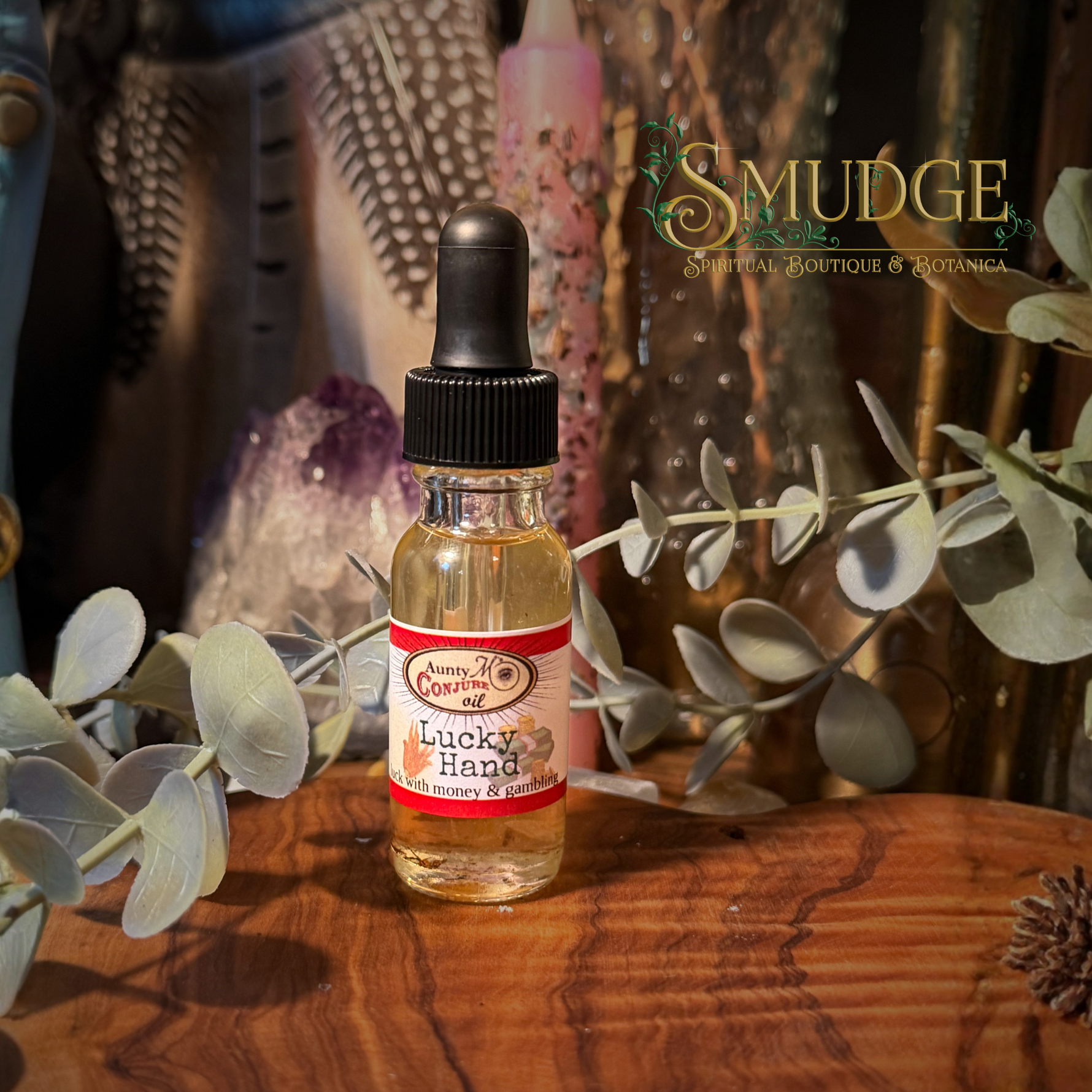 A small glass dropper bottle labeled 'Lucky Hand' by Queen Al's Botanicals sits on a wooden surface with eucalyptus branches and a boho-style decorative background. The brand name 'SMUDGE' appears in the upper right.