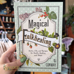 A hand holds a book titled "Magical Folkhealing" by DJ Conway in a shop setting. The book cover has a vintage-style label design with green leaves, red berries, and purple flowers on a pale green and white polka-dotted background.