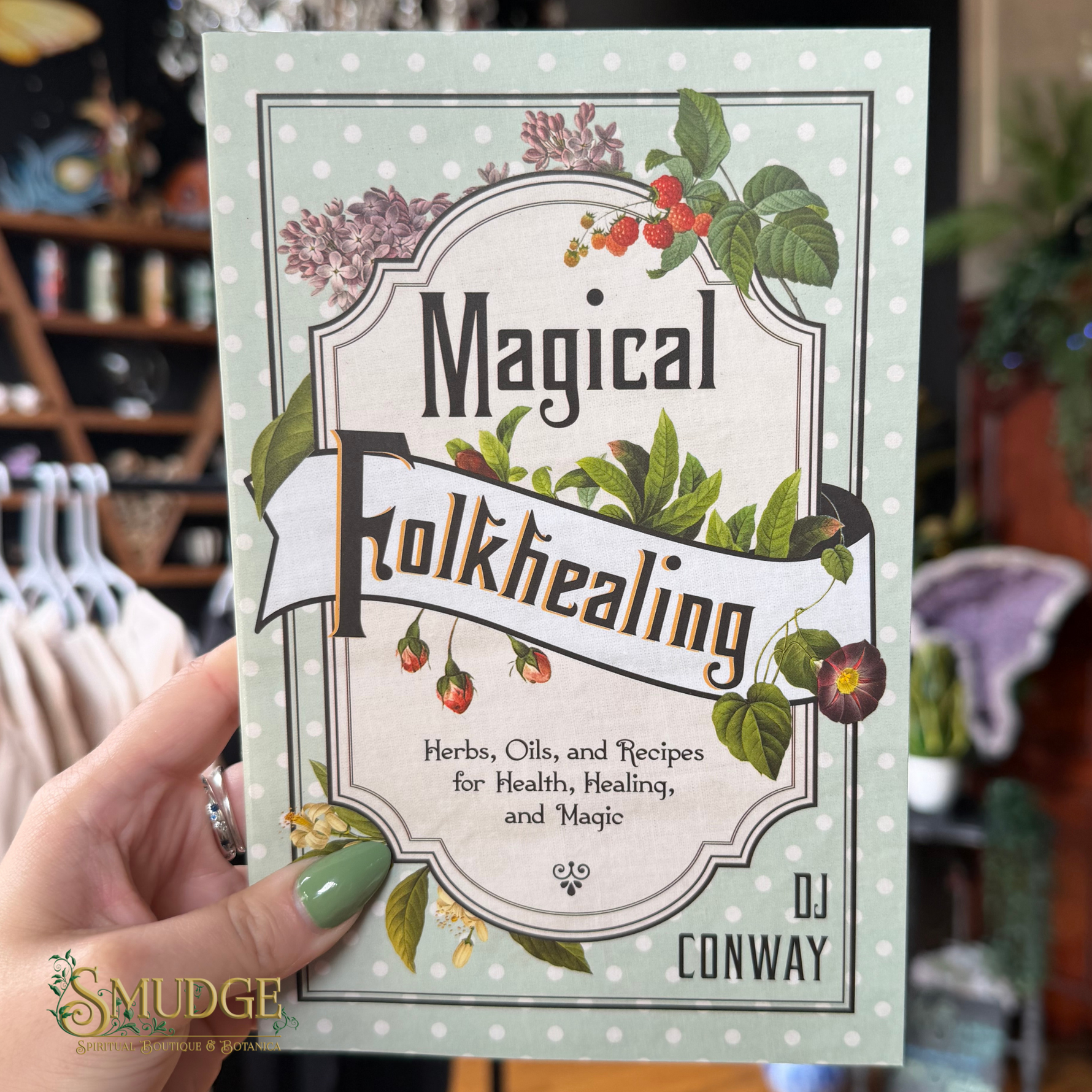 A hand holds a book titled "Magical Folkhealing" by DJ Conway in a shop setting. The book cover has a vintage-style label design with green leaves, red berries, and purple flowers on a pale green and white polka-dotted background.