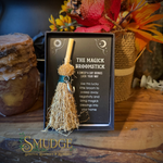 A small broom-topped wand presented in a black gift box with printed instructions, styled on a wooden surface with autumn flowers and candles in the background. The box reads 'THE MAGICK BROOMSTICK' and shows a small natural-fiber broom with a tan handle and dark teal wrap.