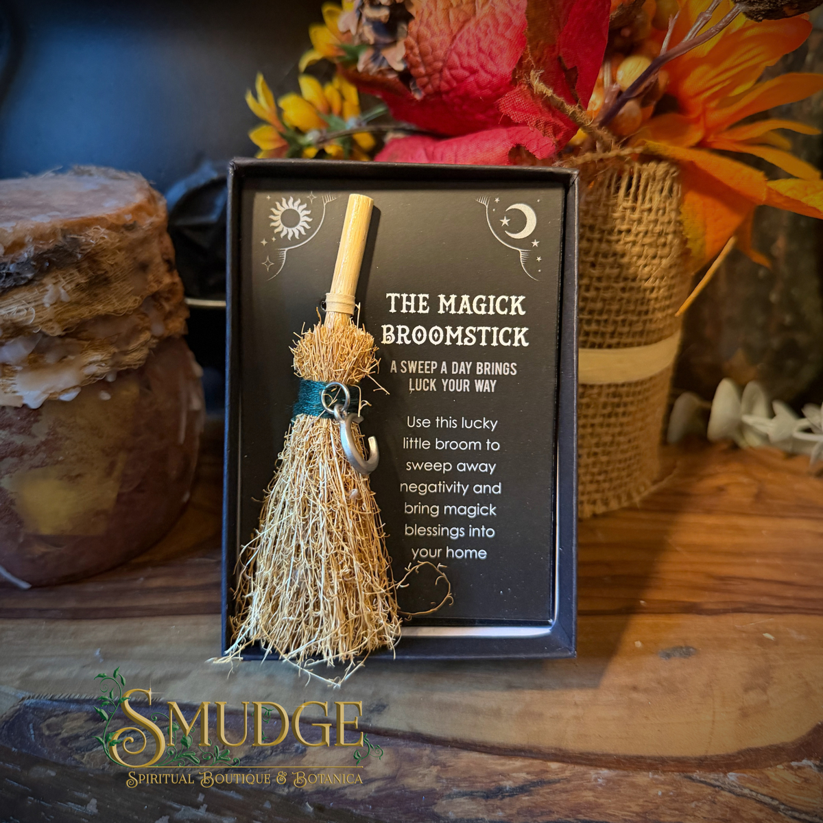 A small broom-topped wand presented in a black gift box with printed instructions, styled on a wooden surface with autumn flowers and candles in the background. The box reads 'THE MAGICK BROOMSTICK' and shows a small natural-fiber broom with a tan handle and dark teal wrap.