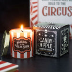 A lit scented candle in a red-and-white striped glass jar sits beside its black-and-white box. The label reads 'Welcome to the Freak Show' and the box reads 'Candy Apple Scented Candle.