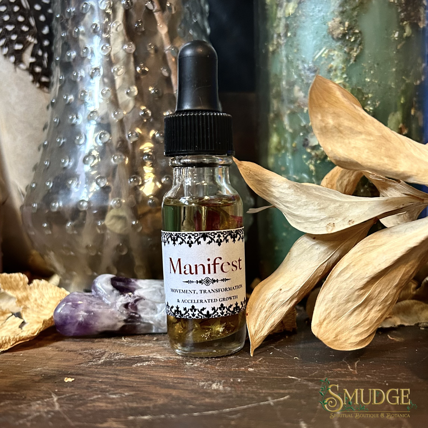 Dropper bottle of Manifest ritual oil on wooden surface that also has foliage and a crystal. 