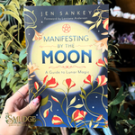 A hand holds a book titled 'Manifesting by the Moon' by Jen Sankey. The book cover features a dark blue background with a large pale yellow moon, blue title text, and red and orange floral illustrations.