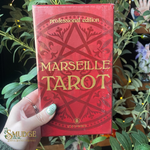Red box of Marseille Tarot with decorative design held by a hand against a green leafy background.