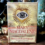 Card deck packaging for 'The Mary Magdalene Oracle' by Meggan Watterson with a heart and eye design sitting on a witchy shelf. 