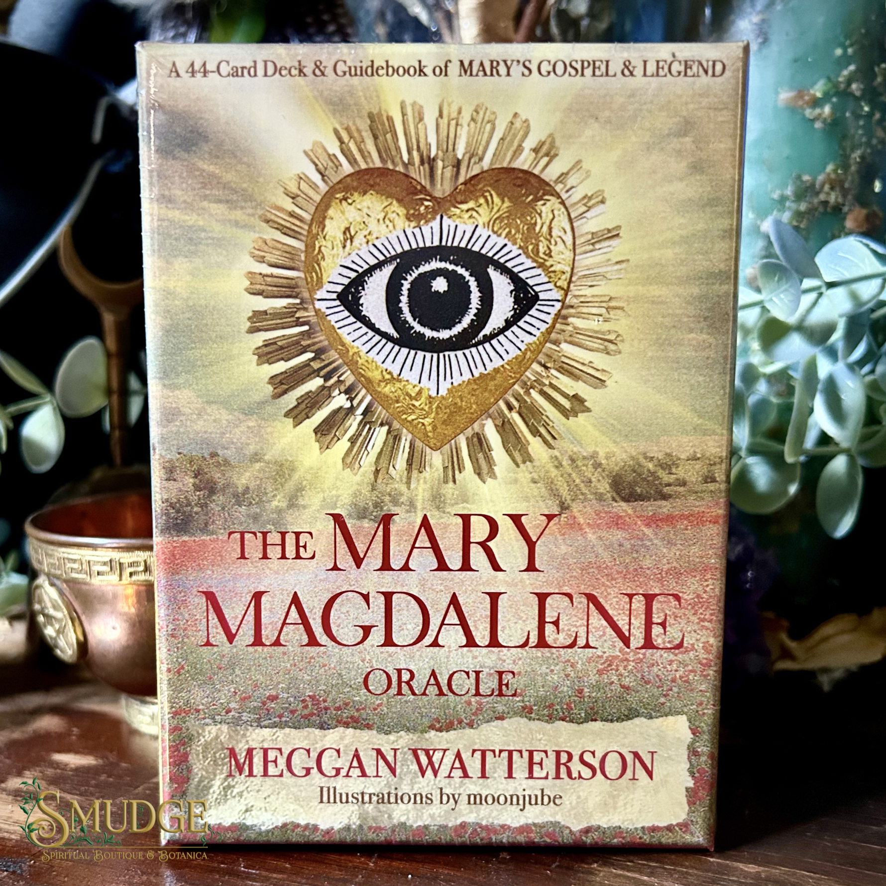 Card deck packaging for 'The Mary Magdalene Oracle' by Meggan Watterson with a heart and eye design sitting on a witchy shelf. 