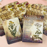 A boxed card deck titled 'Magical Herb Oracle' with its box and two illustrated cards displayed on a soft pink surface, surrounded by botanical artwork and dried floral props.
