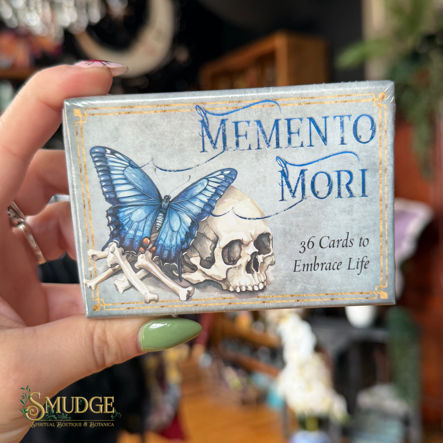 A hand with green nail polish holds a boxed card deck labeled 'MEMENTO MORI' with a blue butterfly, skull, and bones illustration. The box also reads '36 Cards to Embrace Life.