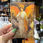 A hand with green nail polish holds a boxed card deck titled "Messages of the Saints" featuring an angel illustration with large golden wings. The background shows a blurred retail shop interior with clothing racks and flowers.