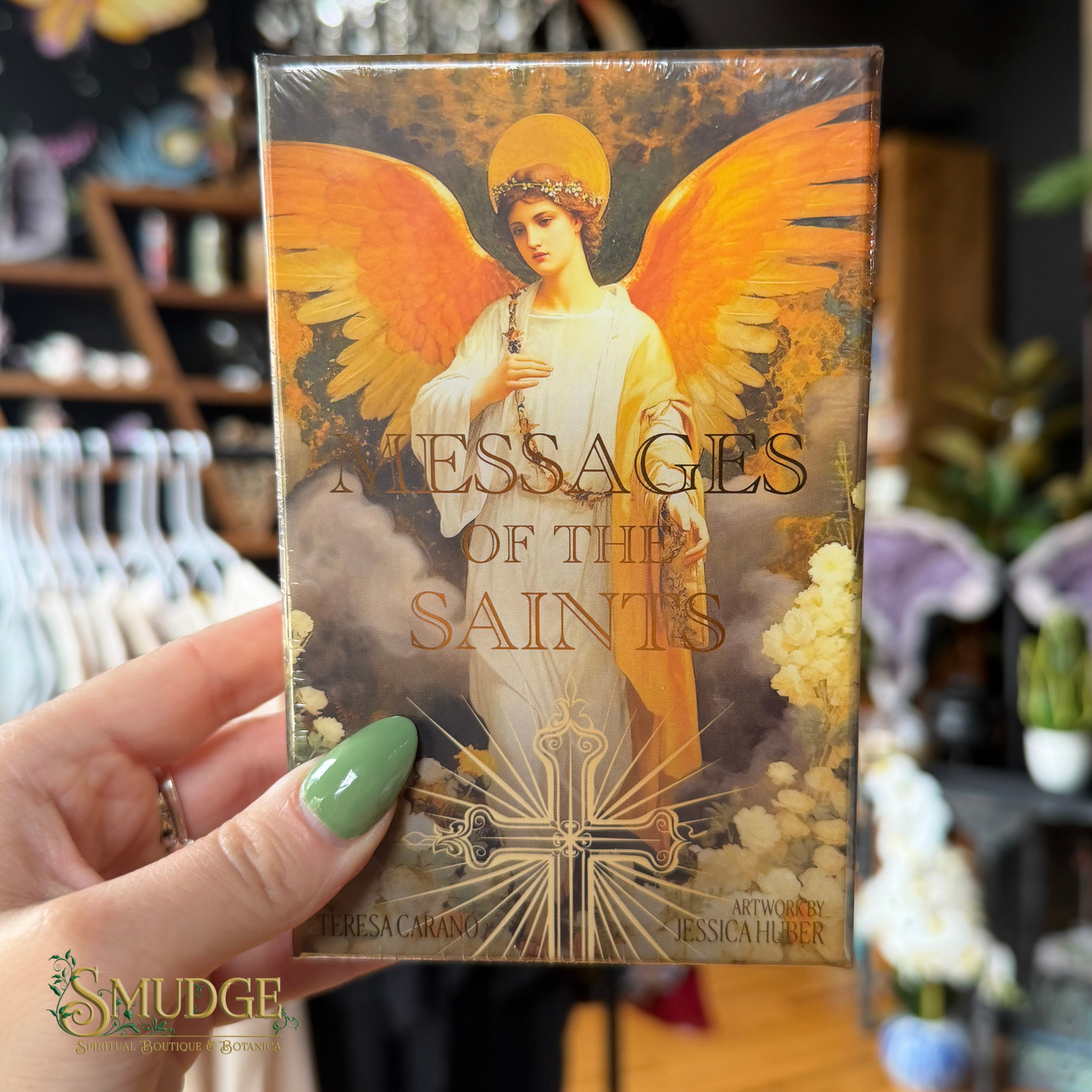 A hand with green nail polish holds a boxed card deck titled "Messages of the Saints" featuring an angel illustration with large golden wings. The background shows a blurred retail shop interior with clothing racks and flowers.