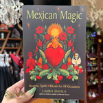 A hand holds a book titled "Mexican Magic" by Laura Davila. The cover features a dark gray background with a colorful central illustration of a red heart, green cacti, red roses, and small skeleton figures.