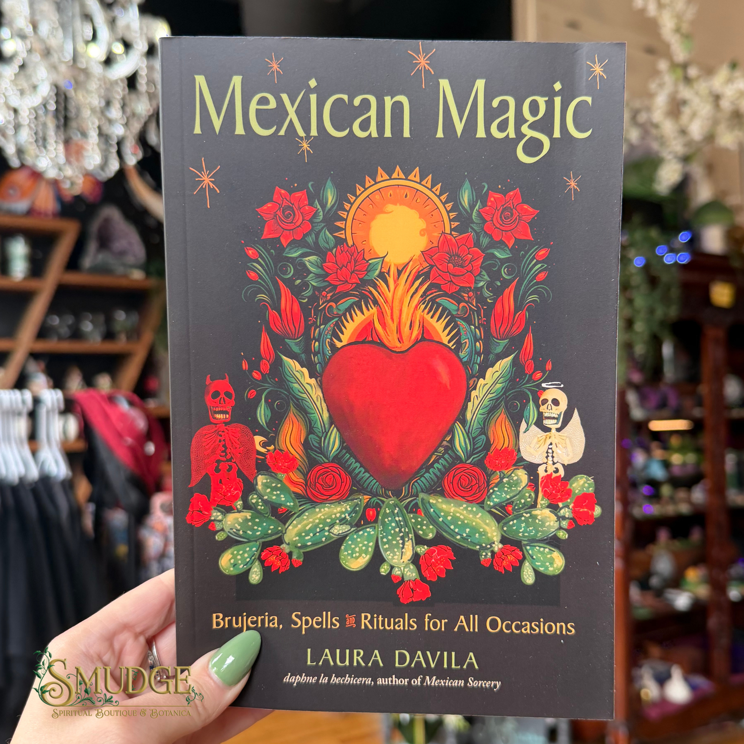 A hand holds a book titled "Mexican Magic" by Laura Davila. The cover features a dark gray background with a colorful central illustration of a red heart, green cacti, red roses, and small skeleton figures.