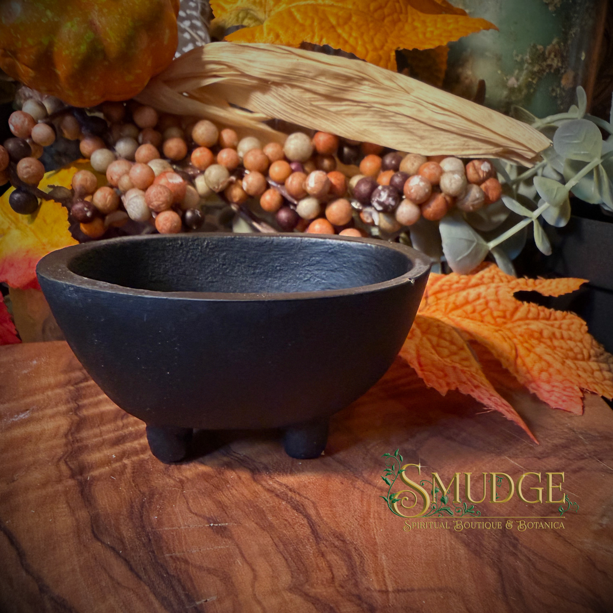 A small black textured ceramic bowl with three feet, displayed on a wooden surface with autumn leaves and decorative beads in the background. The brand logo 'SMUDGE' is visible in the bottom right corner.