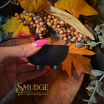 A hand with bright pink fingernails holds a small black footed bowl, set against an autumn-themed background with fall leaves, berries, and a wooden surface. The brand name 'SMUDGE' is visible in the lower-left corner.