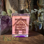 A square glass perfume bottle labeled 'PERFUME MONEY HOUSE BLESSING' with purple liquid and a black cap, styled on a wooden surface with crystals, dried leaves, and greenery in the background. A 'Smudge' brand watermark is visible at the bottom left.
