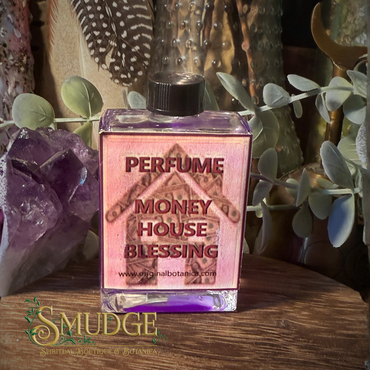 A square glass perfume bottle labeled 'PERFUME MONEY HOUSE BLESSING' with purple liquid and a black cap, styled on a wooden surface with crystals, dried leaves, and greenery in the background. A 'Smudge' brand watermark is visible at the bottom left.
