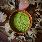 Bowl of green incense powder with a pink candle and flowers on a wooden surface, branded 'Smudge'.