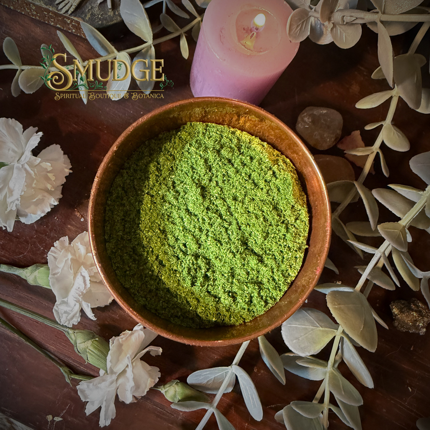 Bowl of green incense powder with a pink candle and flowers on a wooden surface, branded 'Smudge'.