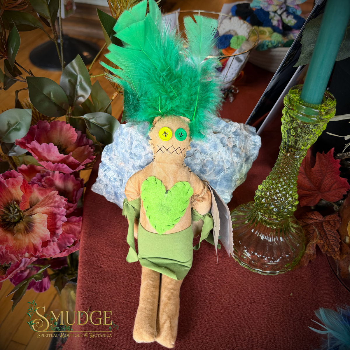 A handmade rag doll with a bright green feathered wig, a green heart-shaped chest piece, and a green skirt, posed on a red tablecloth surrounded by flowers, a glass candle holder, and a blue feather.