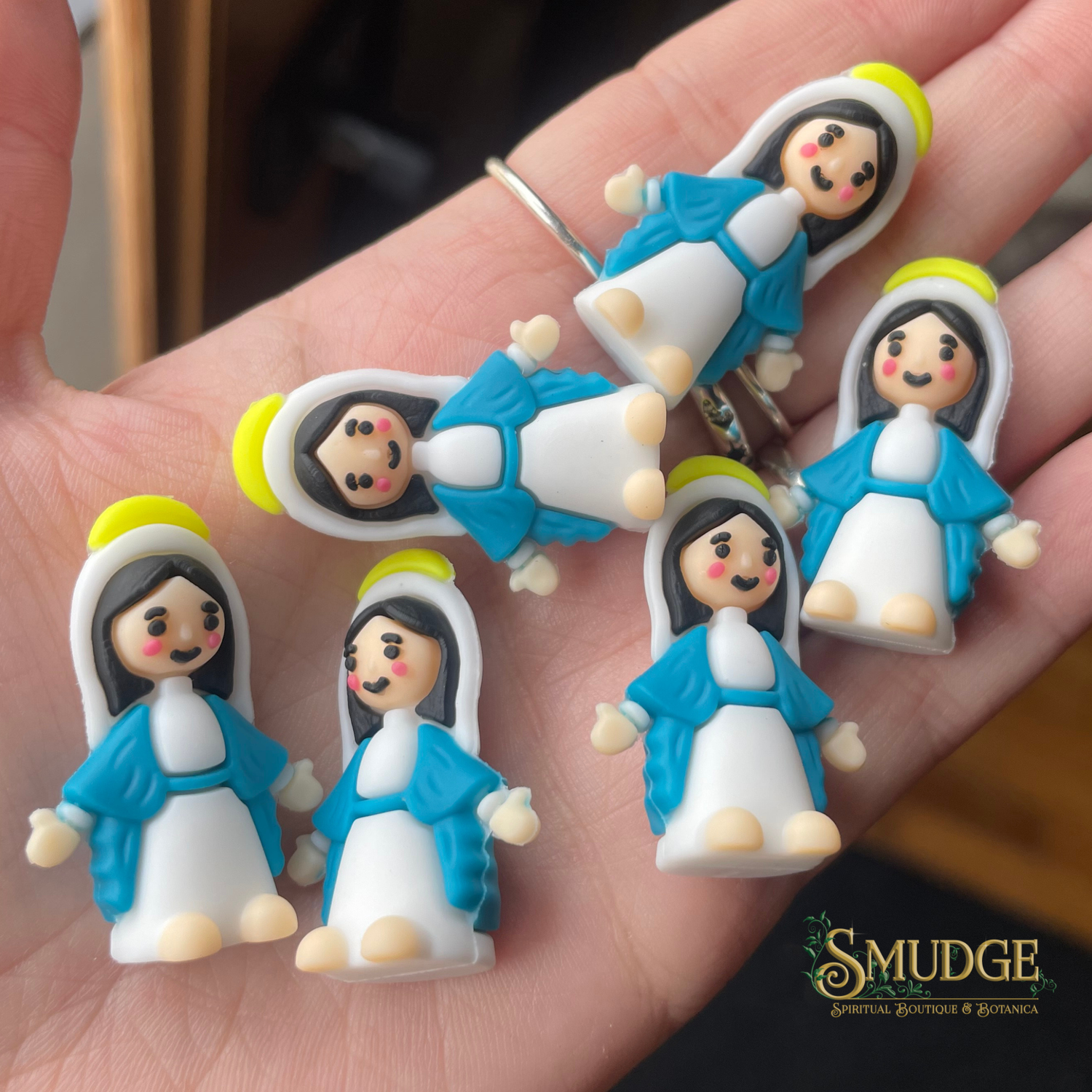 Small figurines of a woman in blue and white dress (Mother Mary) on a hand, with 'Smudge' branding.