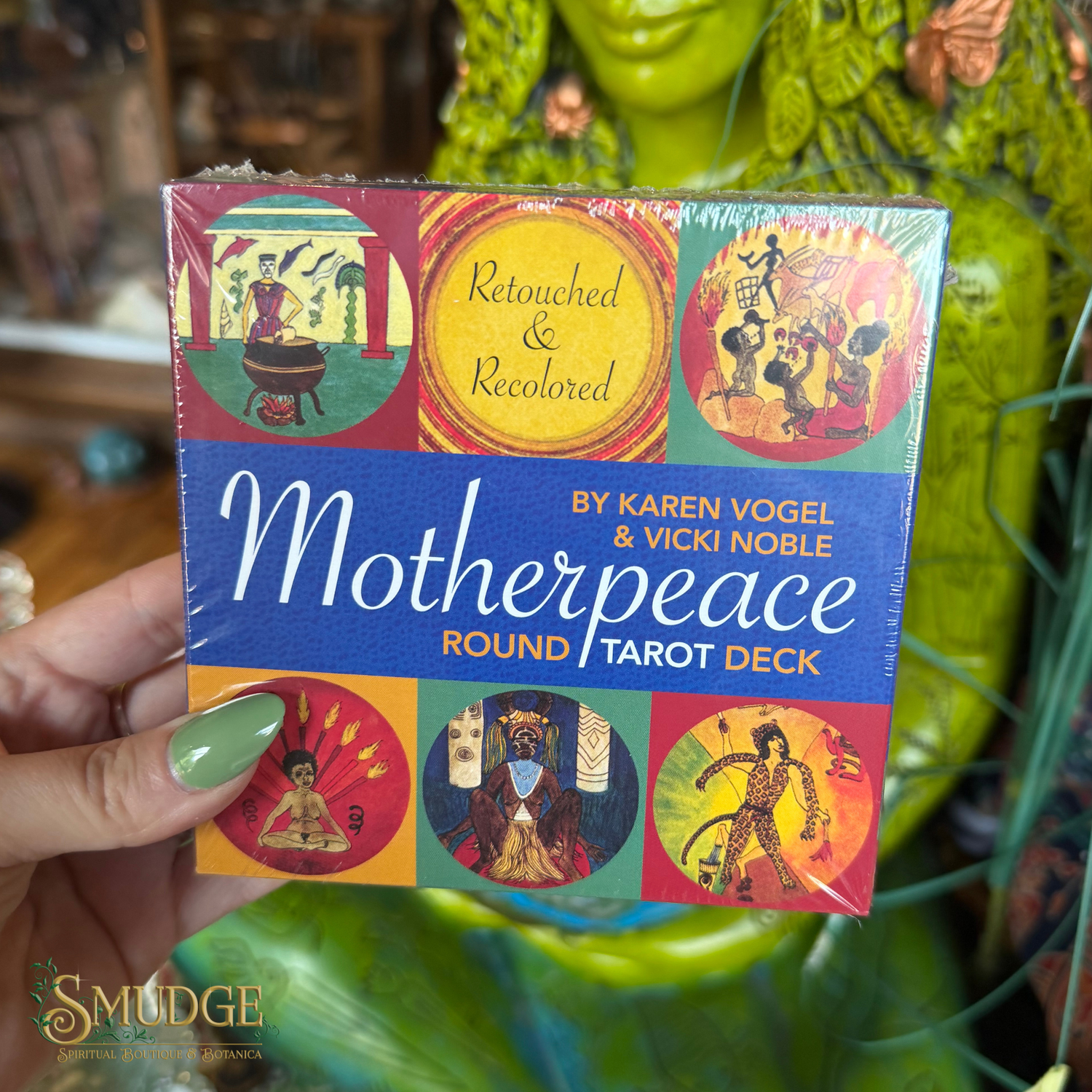 A hand with green nail polish holds a shrink-wrapped copy of the Motherpeace Tarot Deck. The box features a blue center band with white script text and colorful circular illustrations around it.