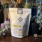 A stand-up pouch of Smudge Apothecary Wellness Tea, specifically Mullein Leaf, displayed on a wooden surface surrounded by dried botanicals, a candle, and glass jars.