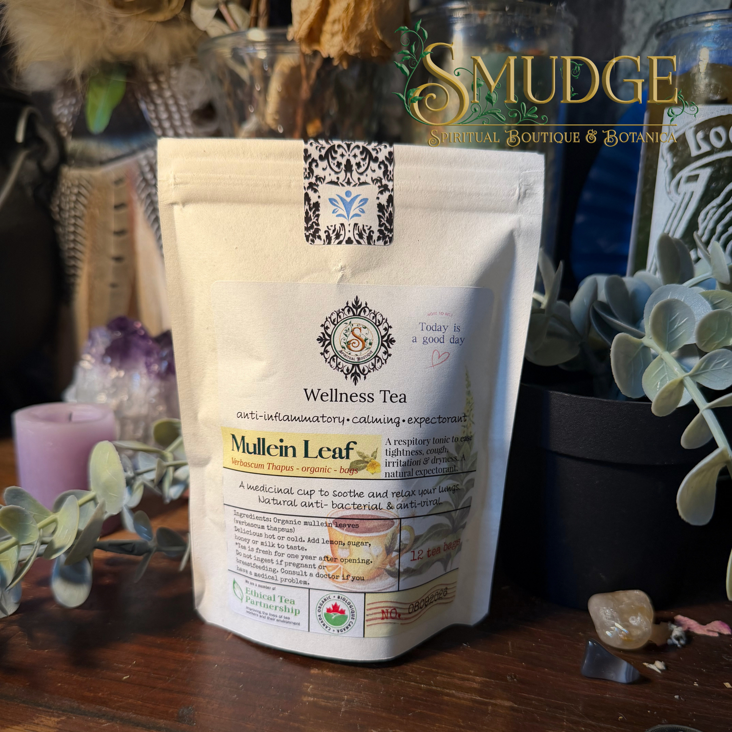 A stand-up pouch of Smudge Apothecary Wellness Tea, specifically Mullein Leaf, displayed on a wooden surface surrounded by dried botanicals, a candle, and glass jars.