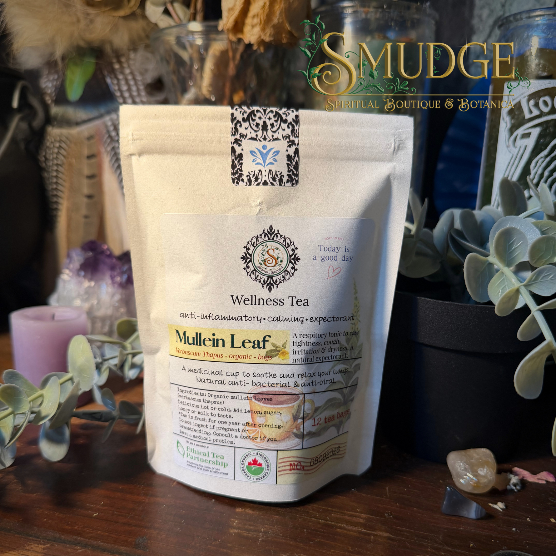 A stand-up pouch of Smudge Apothecary Wellness Tea, specifically Mullein Leaf, displayed on a wooden surface surrounded by dried botanicals, a candle, and glass jars.