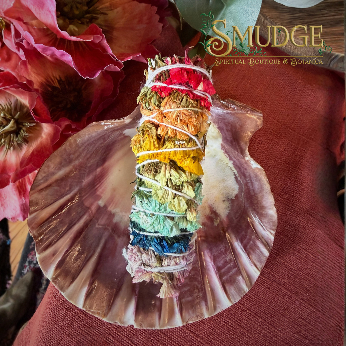 A rainbow-colored herbal smudge bundle wrapped in white string, displayed on a large clam shell with pink flowers and a rust-red fabric background. The brand logo 'SMUDGE' is visible in the top right corner.