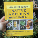 Book titled 'A Beginner's Guide to Native American Herbal Medicine' held by a hand with green nail polish, surrounded by plants.