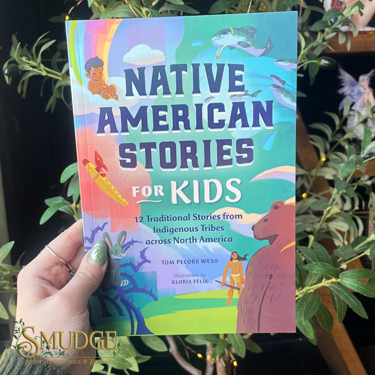 Book titled 'Native American Stories for Kids' held by a hand with green nail polish, surrounded by plants.