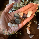 A hand holds a small collection of polished tumbled stones in various colors, including gray, cream, purple, and brown, with more stones and decorative plants in the background on a wooden surface. A logo at the bottom left reads 'SMUDGE Spiritual Boutique & Botanica'.
