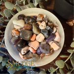 A collection of polished tumbled gemstones in various colors, including purple, pink, orange, brown, and gray, arranged in a white ceramic bowl. The bowl is placed on a dark wooden surface and surrounded by green eucalyptus leaves.