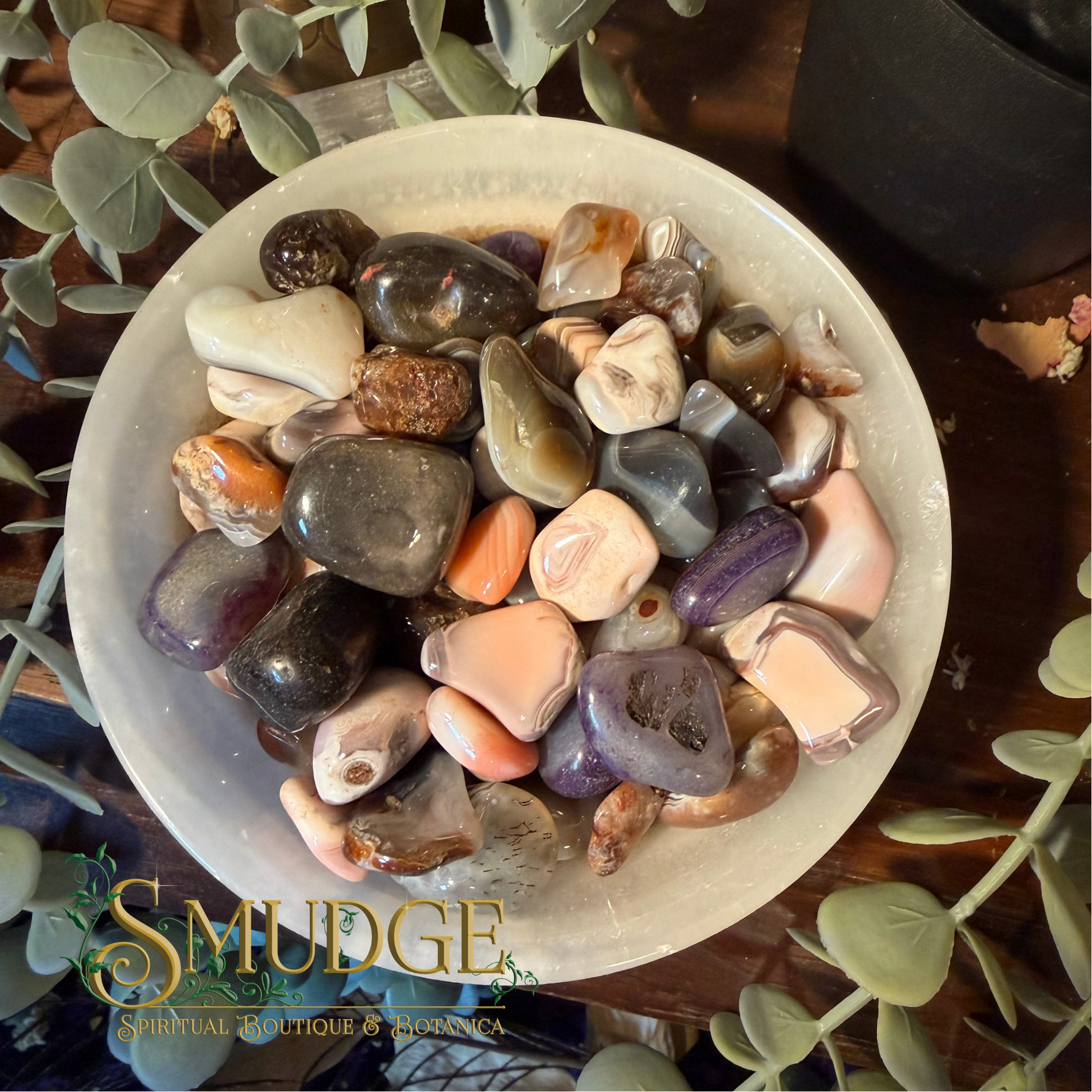 A collection of polished tumbled gemstones in various colors, including purple, pink, orange, brown, and gray, arranged in a white ceramic bowl. The bowl is placed on a dark wooden surface and surrounded by green eucalyptus leaves.