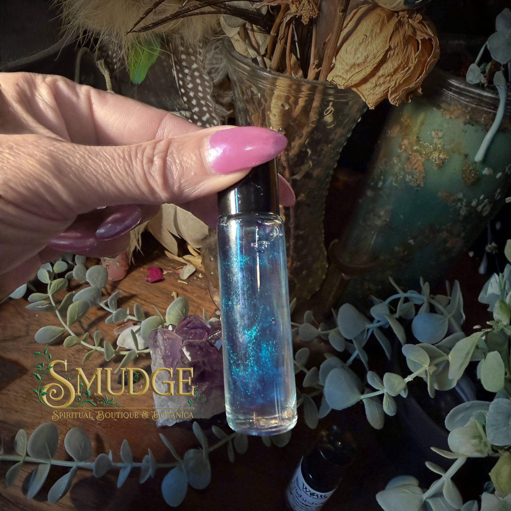 A hand with pink nails holds a clear glass roller bottle filled with shimmering blue liquid. The background is a styled tabletop with dried flowers, greenery, crystals, and a visible 'Smudge Spiritual Boutique & Supply' logo watermark.