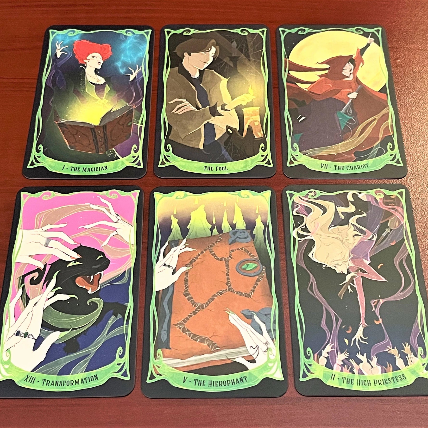 Six illustrated oracle cards arranged in two rows on a reddish-brown wooden surface. The cards feature fantasy artwork with green borders and titles including The Magician, The Fool, The Chariot, The Transformation, The Hermit, and The High Priestess.