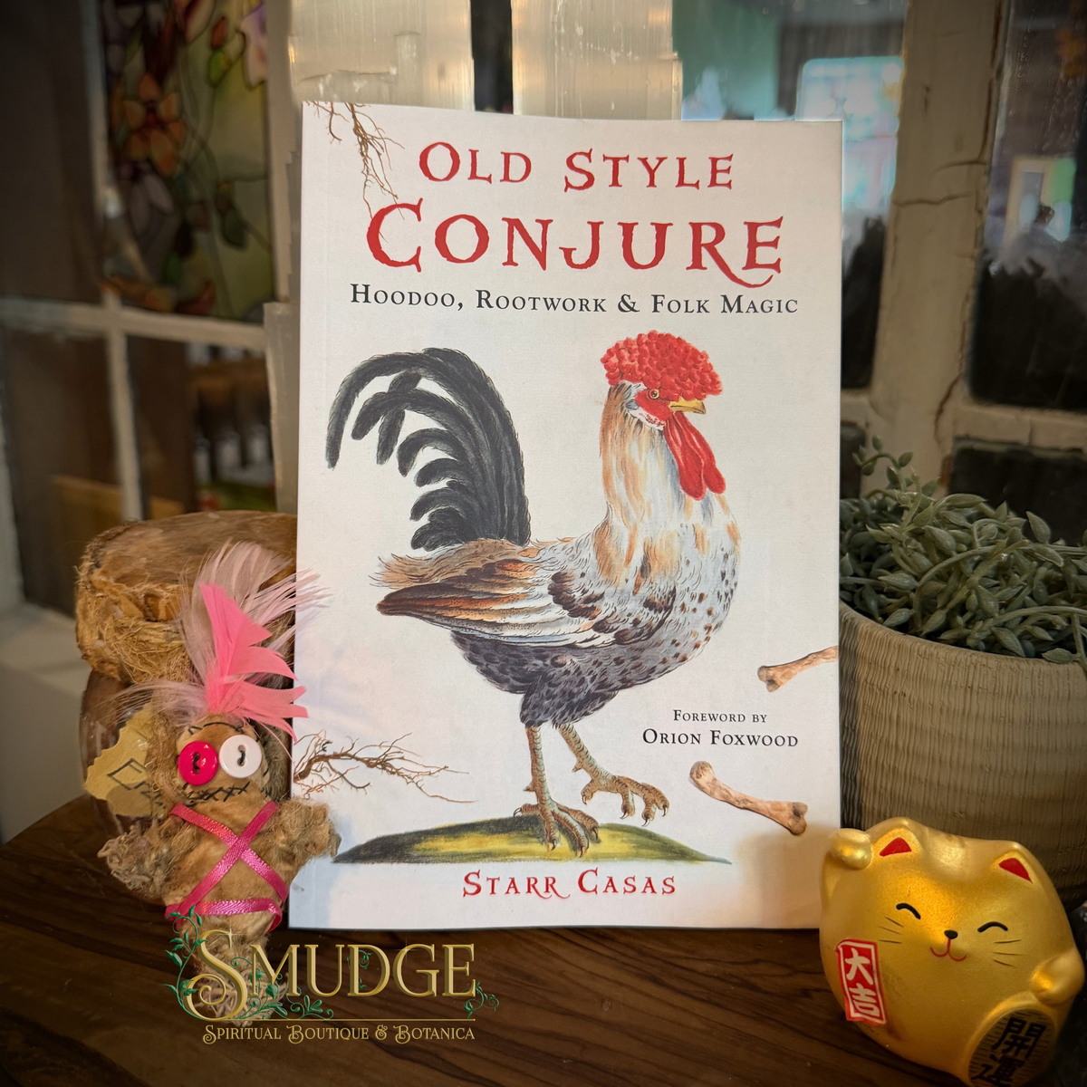 The book 'Old Style Conjure' by Starr Casas is displayed upright on a wooden surface, flanked by a small owl figurine with pink feathers and a gold lucky cat statue. The book cover features a detailed illustration of a rooster.