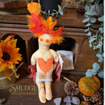 A handmade autumn-themed rag doll with orange and yellow feathered hair, a stitched orange heart on its chest, and a plaid skirt, styled on a rustic wooden shelf with fall decor.