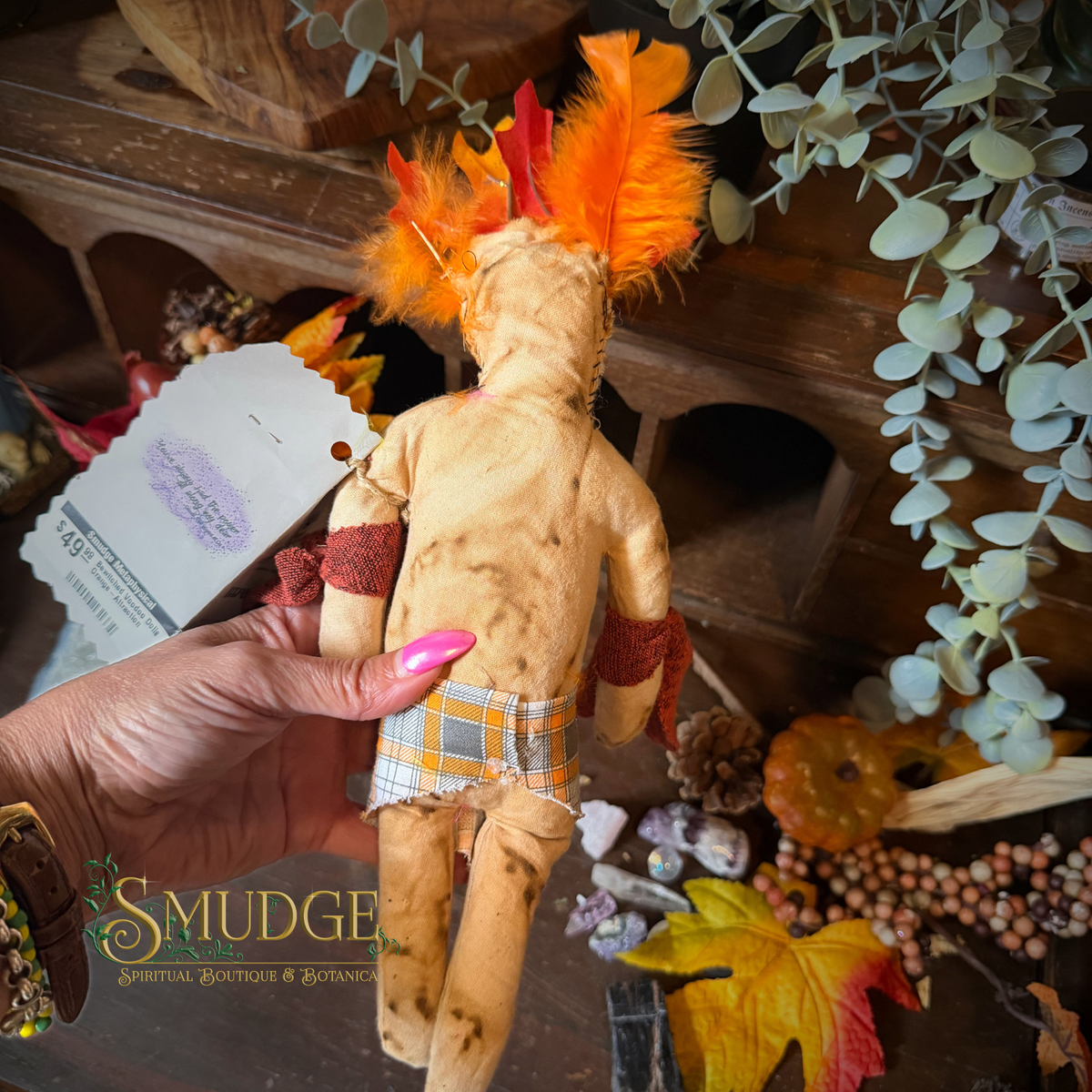 A handmade wooden spiritual doll with orange and red feathers, a plaid skirt, and red arm bands is held by a hand with pink nail polish. The background is a rustic wooden shelf decorated with eucalyptus, autumn leaves, and crystals, with a 'SMUDGE Spiritual Boutiques & Crafts' logo in the bottom-left corner.