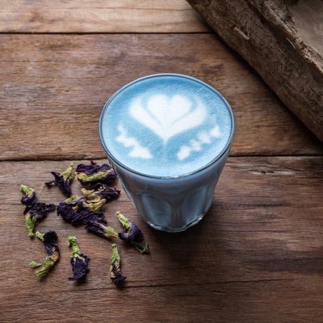 A glass of light blue latte with white heart-shaped latte art sits on a rustic wooden table next to dried purple butterfly pea flowers.