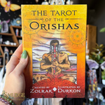 A hand holds a book titled 'The Tarot of the Orishas' by Zolrak Durkon. The book cover features a warm yellow-to-orange gradient background with an illustration of an Orisha seated in the center, wearing red garments and holding a sword.