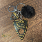 Ouija keychain with a black pom-pom on a wooden surface, branded 'Smudge'.