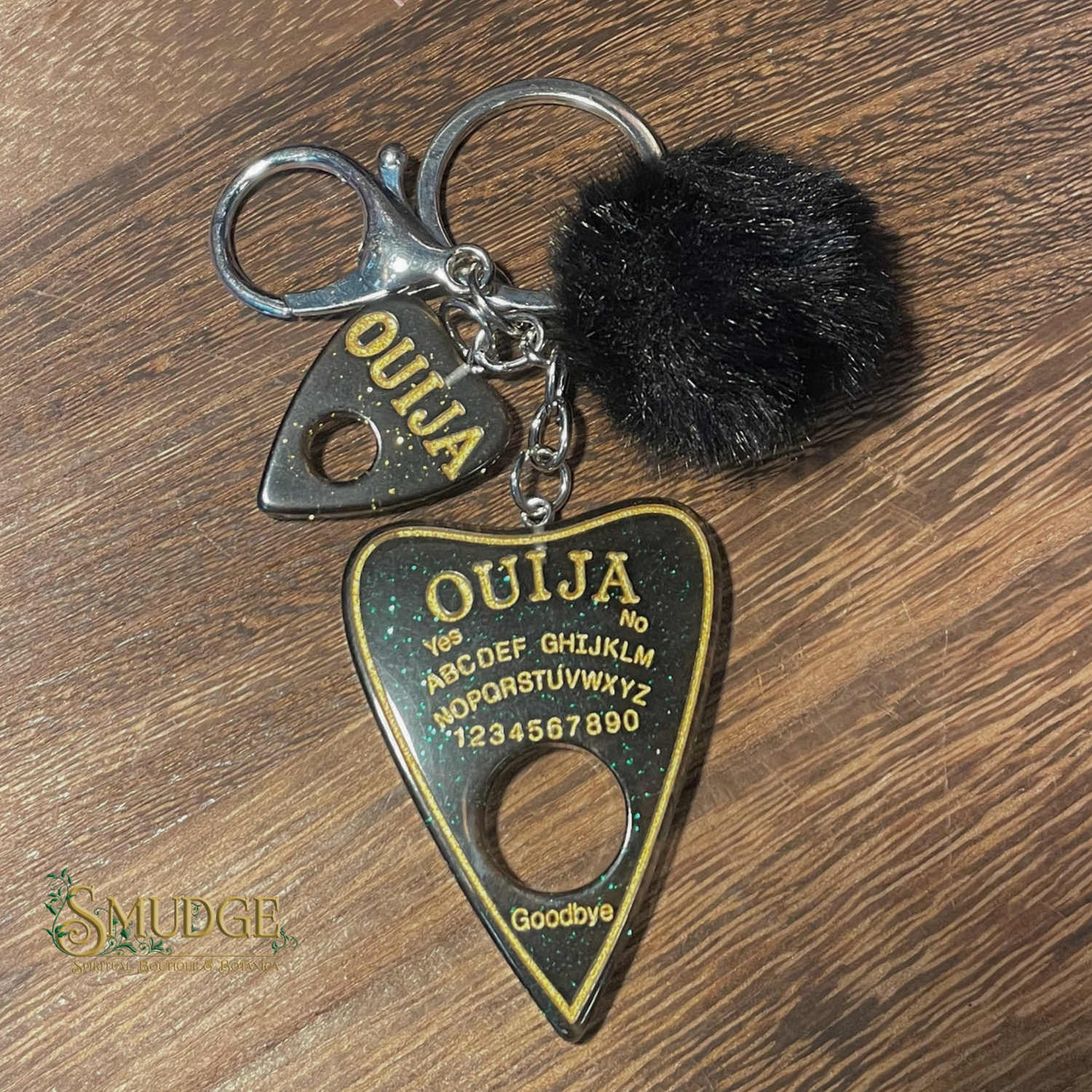 Ouija keychain with a black pom-pom on a wooden surface, branded 'Smudge'.